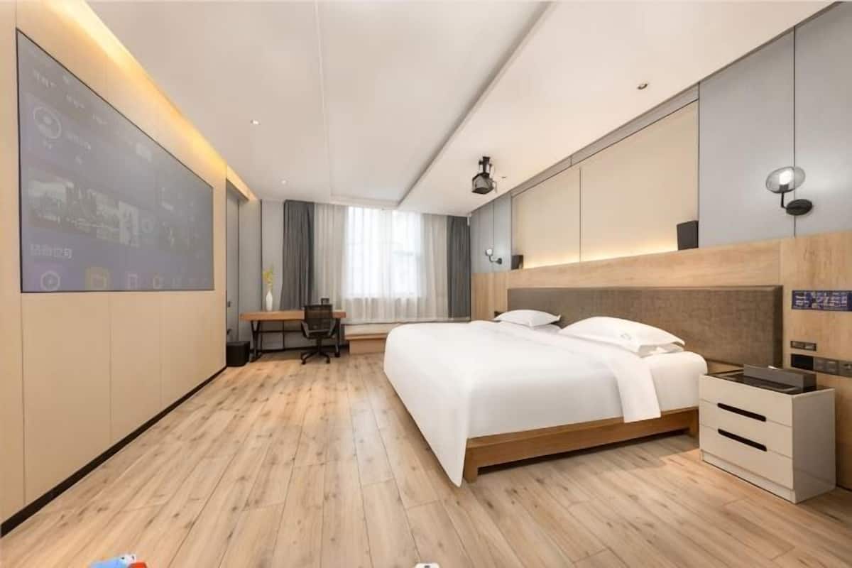 A modern hotel room with a large bed, a desk, and a TV screen on the wall.