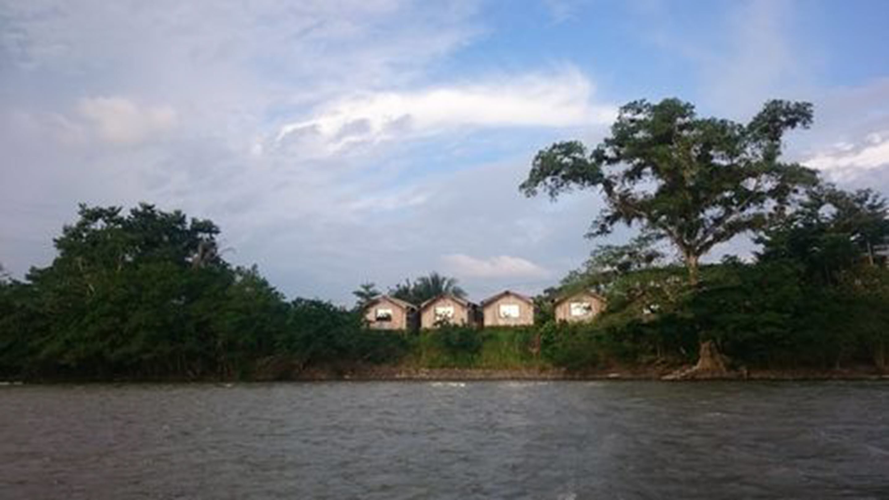 Une rangée de trois petits bâtiments à un seul étage, dotés d’un toit plat, entourés d’une végétation luxuriante et avec une étendue d’eau au premier plan.