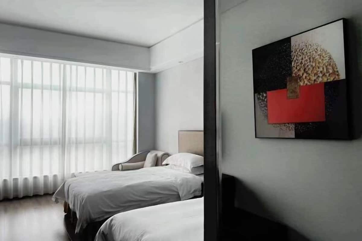 A modern hotel room with two beds, a large window with sheer curtains, and a contemporary abstract painting on the wall.