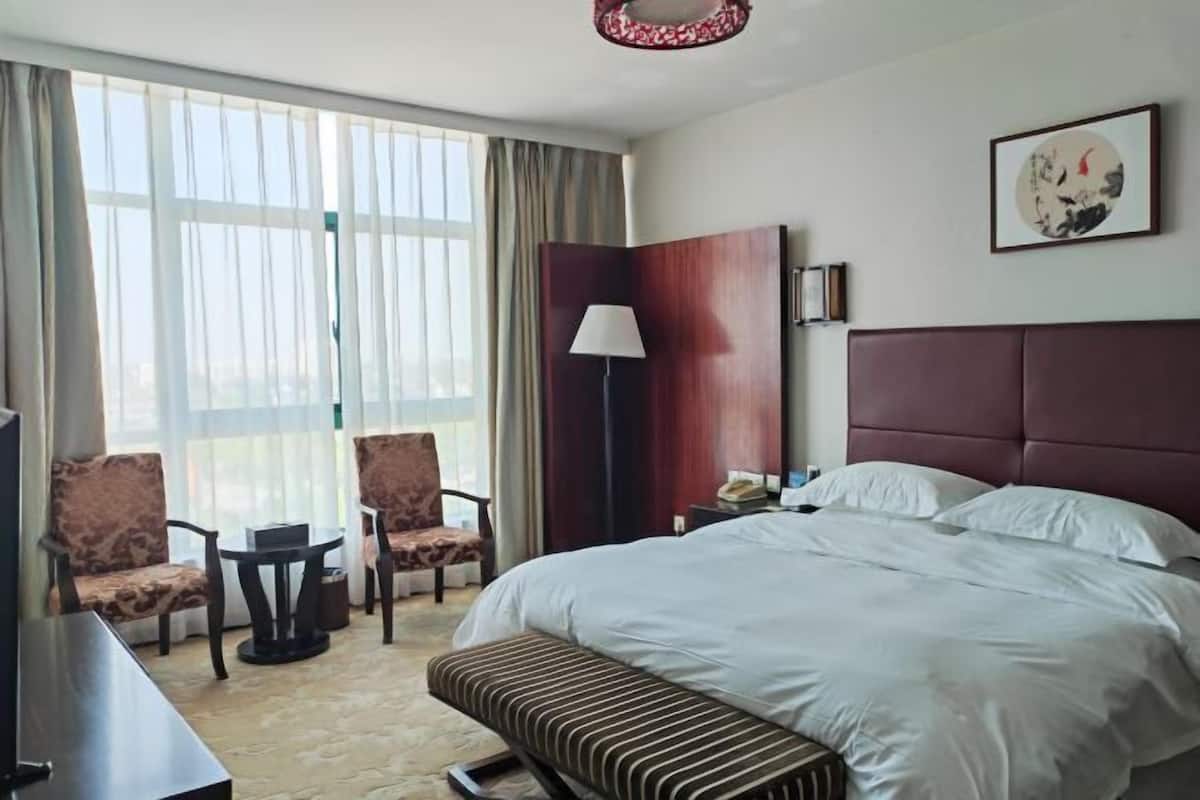 A hotel room with a large bed, a desk, two chairs, a small table, a lamp, and a large window with curtains.