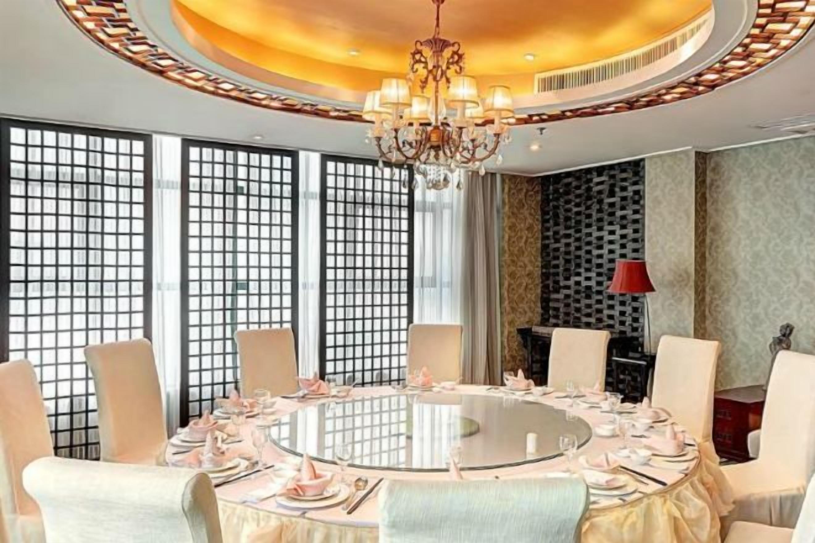 A round dining table with chairs, a chandelier, and a view through a window with a grid pattern.