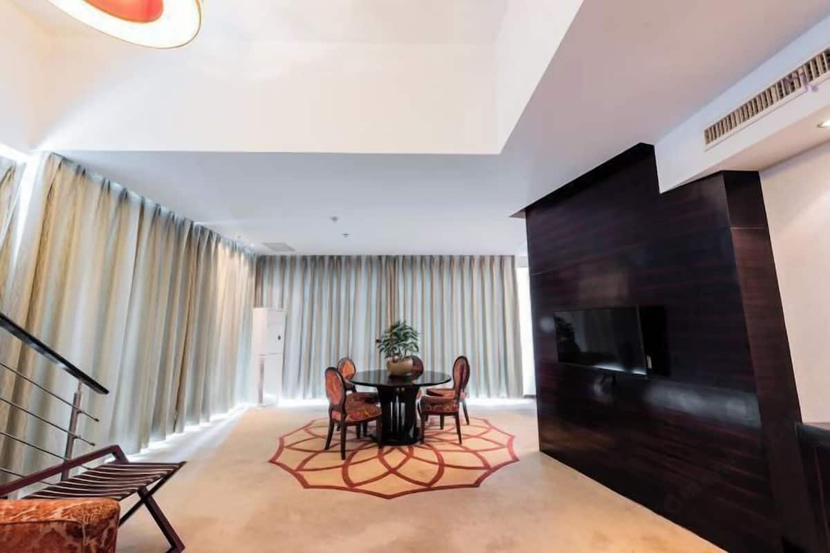 A modern hotel room with a round dining table, four chairs, a potted plant, a large flat-screen TV, and a dark wood accent wall.