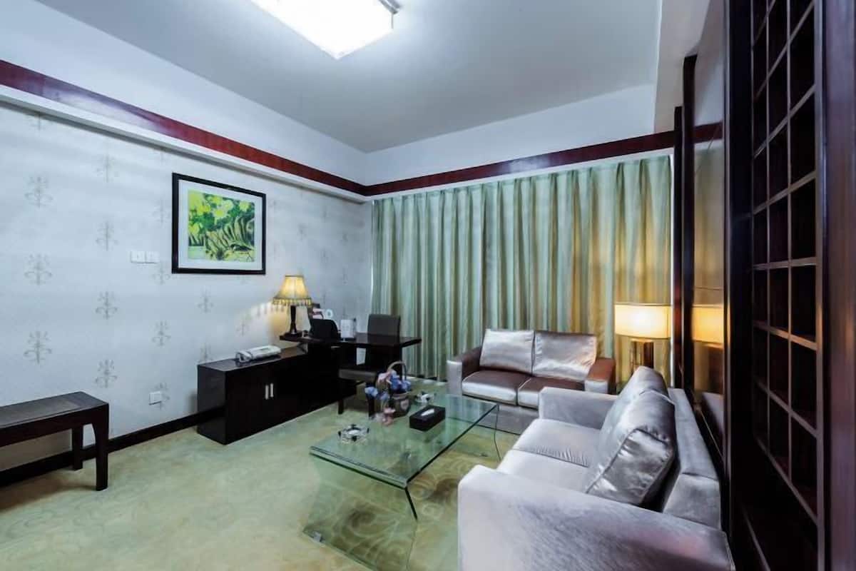 A living room with a sofa, a glass coffee table, a desk, and a chair. There is a painting on the wall and a window with curtains.