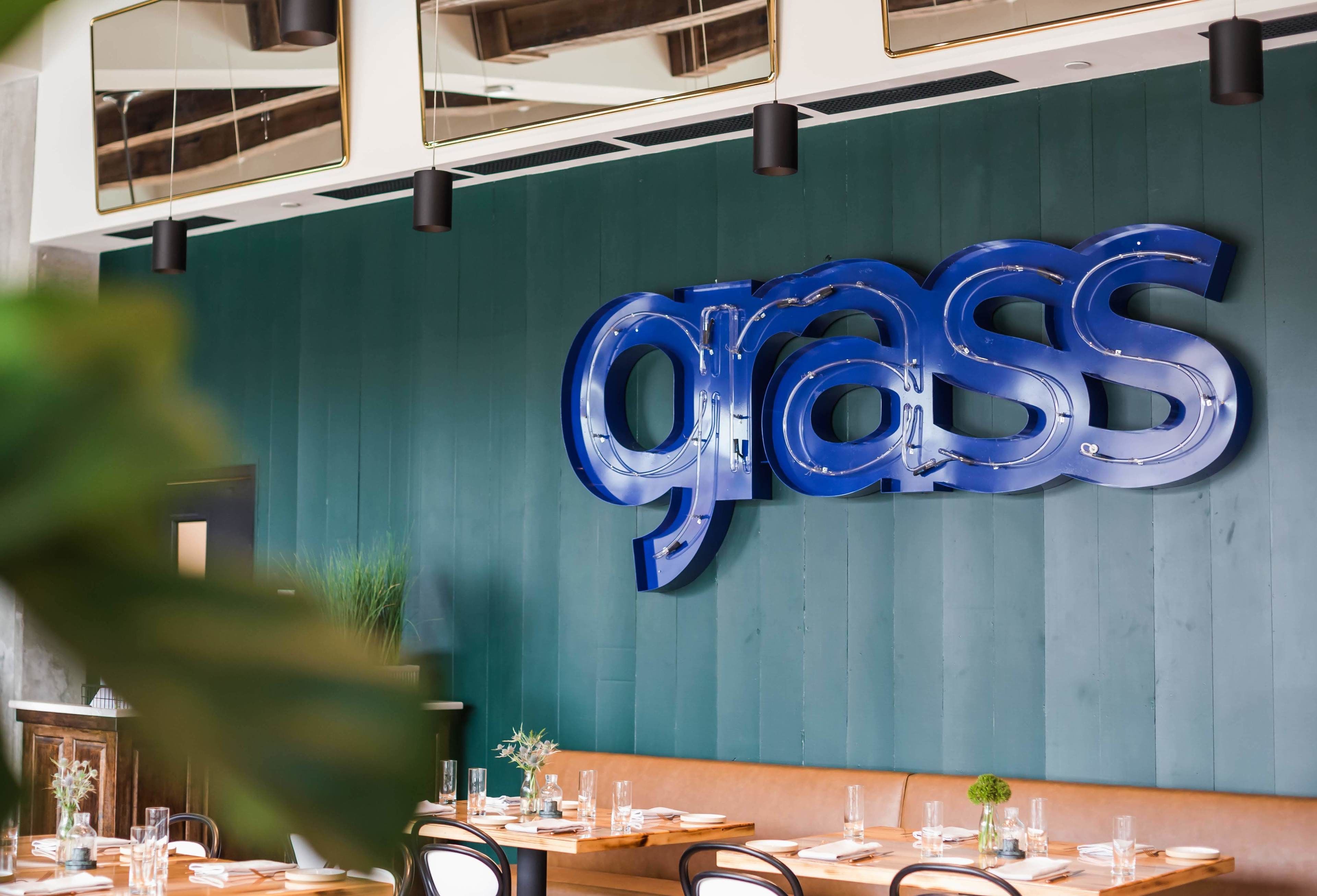 Ein Restaurantinnenraum mit einer türkisfarbenen Wand und einem großen, blauen 'grass'-Schild. Die Tische sind mit weißen Tischdecken und Gläsern gedeckt.
