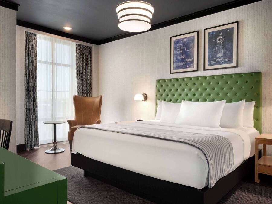 Et hotelværelse med en stor seng, et grønt polstret sengegærde, et lille bord, en stol og to billeder i rammer på væggen.