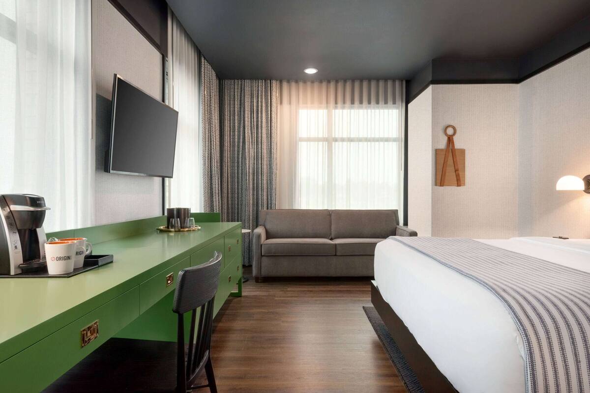A hotel room with a green desk, a flat-screen TV, a grey sofa, a bed with a striped blanket, and a window with curtains.