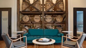 A tasting room with a teal sofa, wooden chairs, and a table with books. Barrels are displayed in the background. - Origin Lexington a Wyndham Hotel (Lexington)