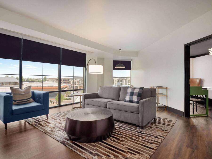 A modern living room with a sofa, armchair, and round coffee table. Large windows offer a view of the city.