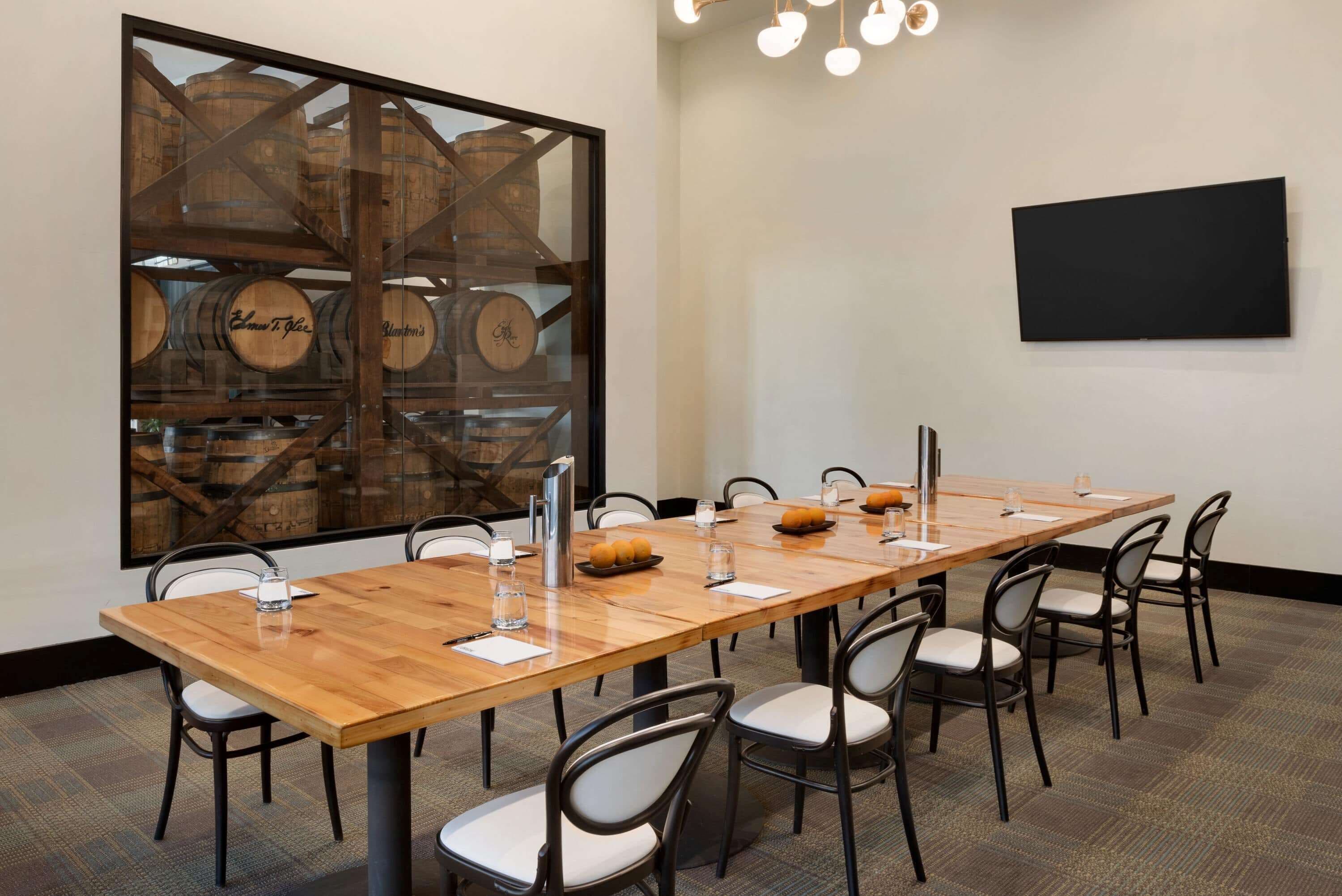 A conference room with a long wooden table, black chairs, a large window with wooden barrels, and a flat-screen TV on the wall.