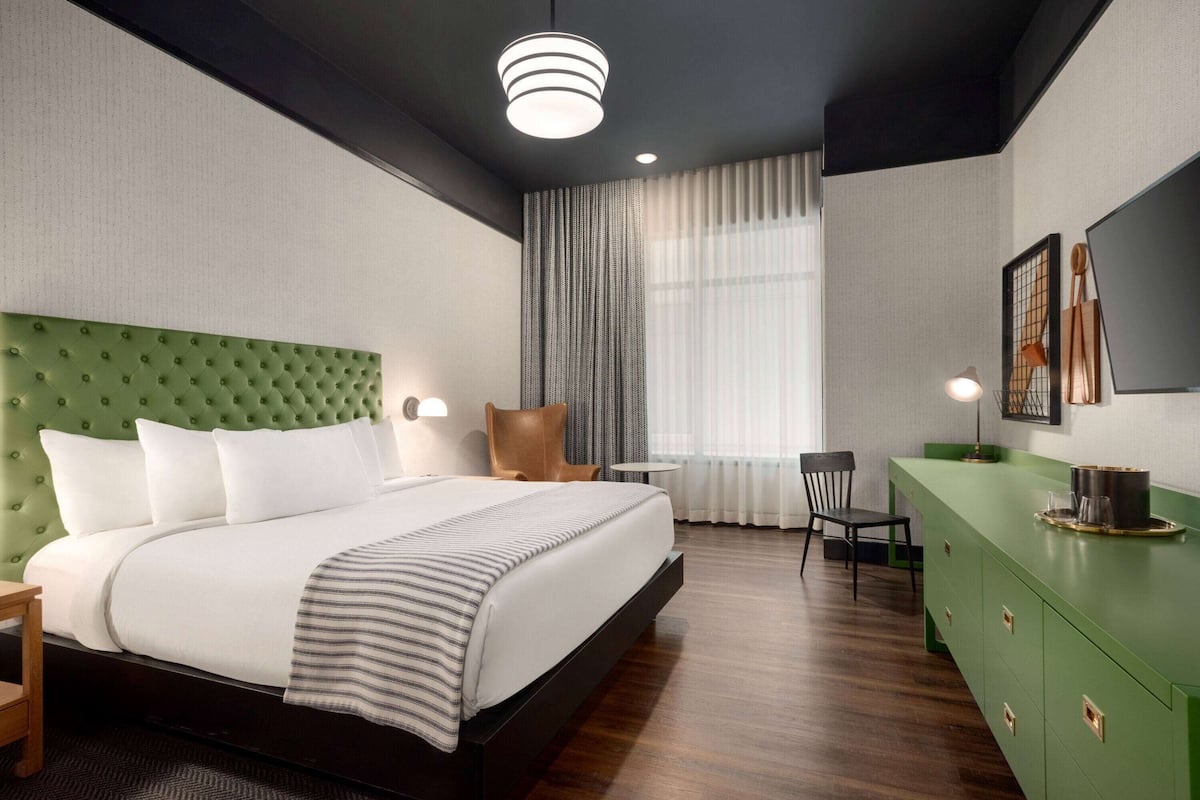 A modern hotel room with a green upholstered headboard, a large bed, a desk with a chair, a green dresser, and a large window with curtains.