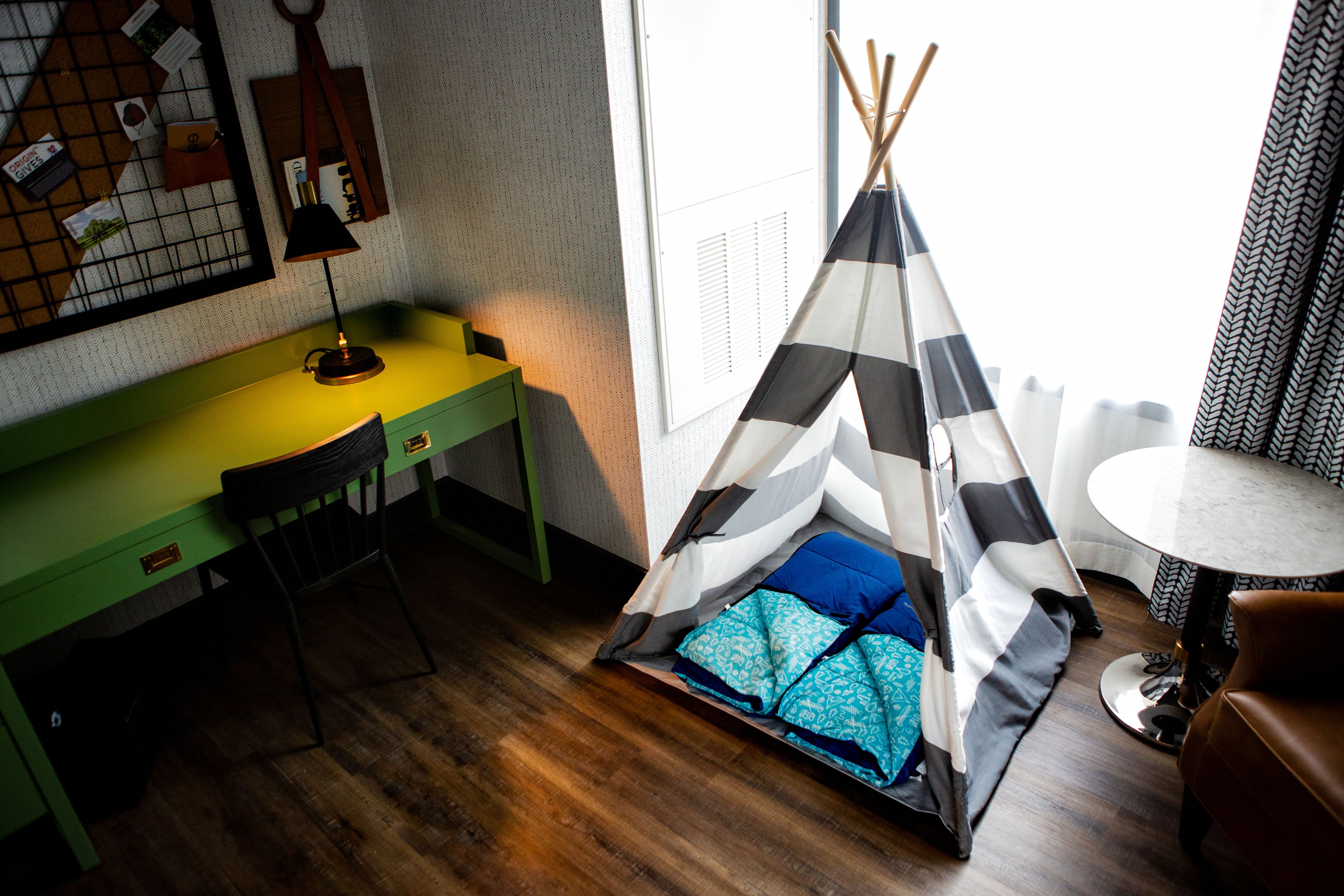 A room with a green desk, a chair, a small table, a teepee with pillows, and a window with blinds.