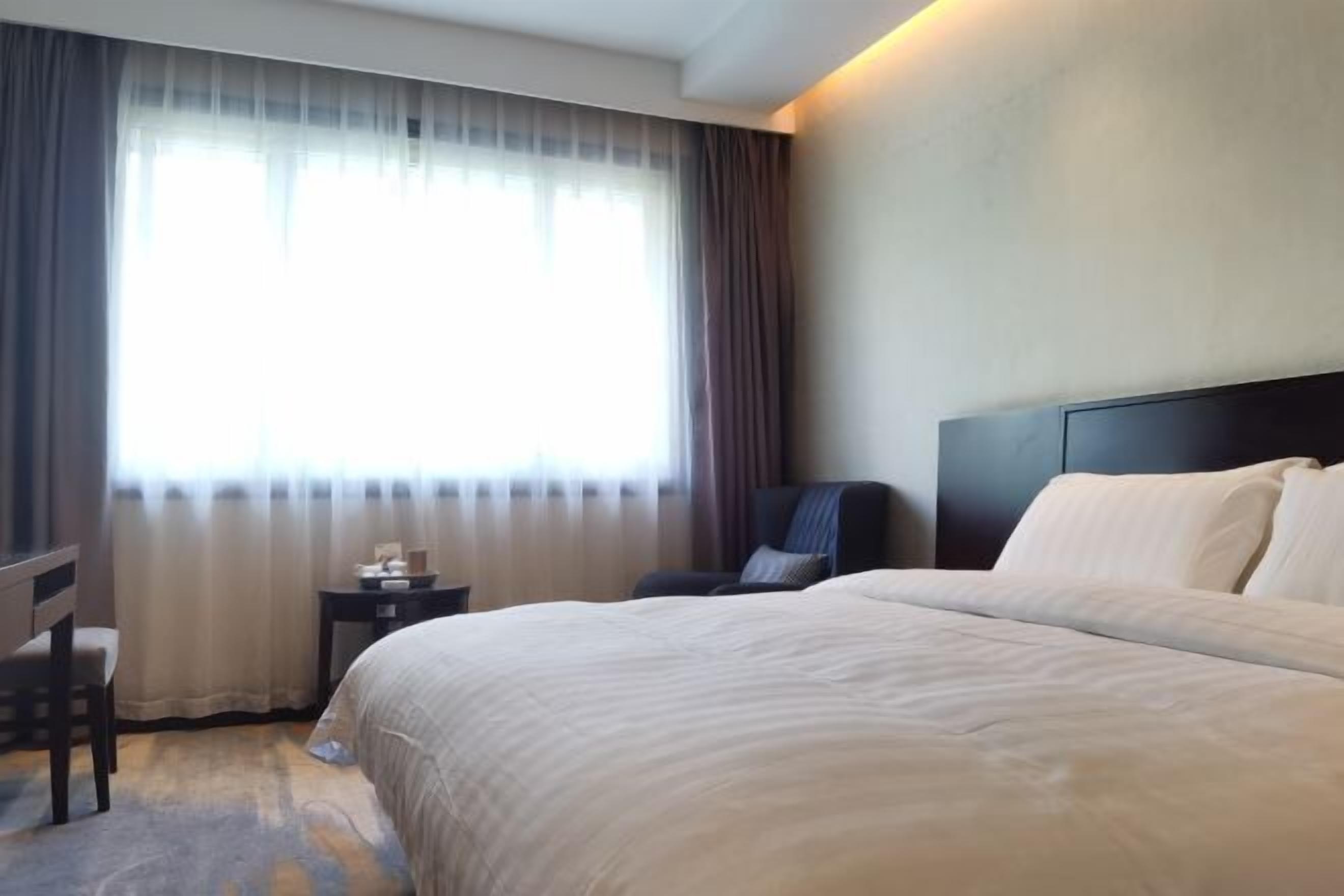 Quarto de hotel com uma cama grande, uma escrivaninha, uma cadeira, uma mesinha e uma janela com cortinas.