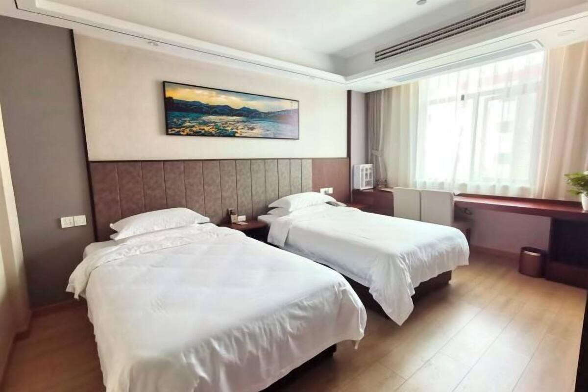 A hotel room with two beds, a painting on the wall, a window with curtains, and a desk with a trash can.
