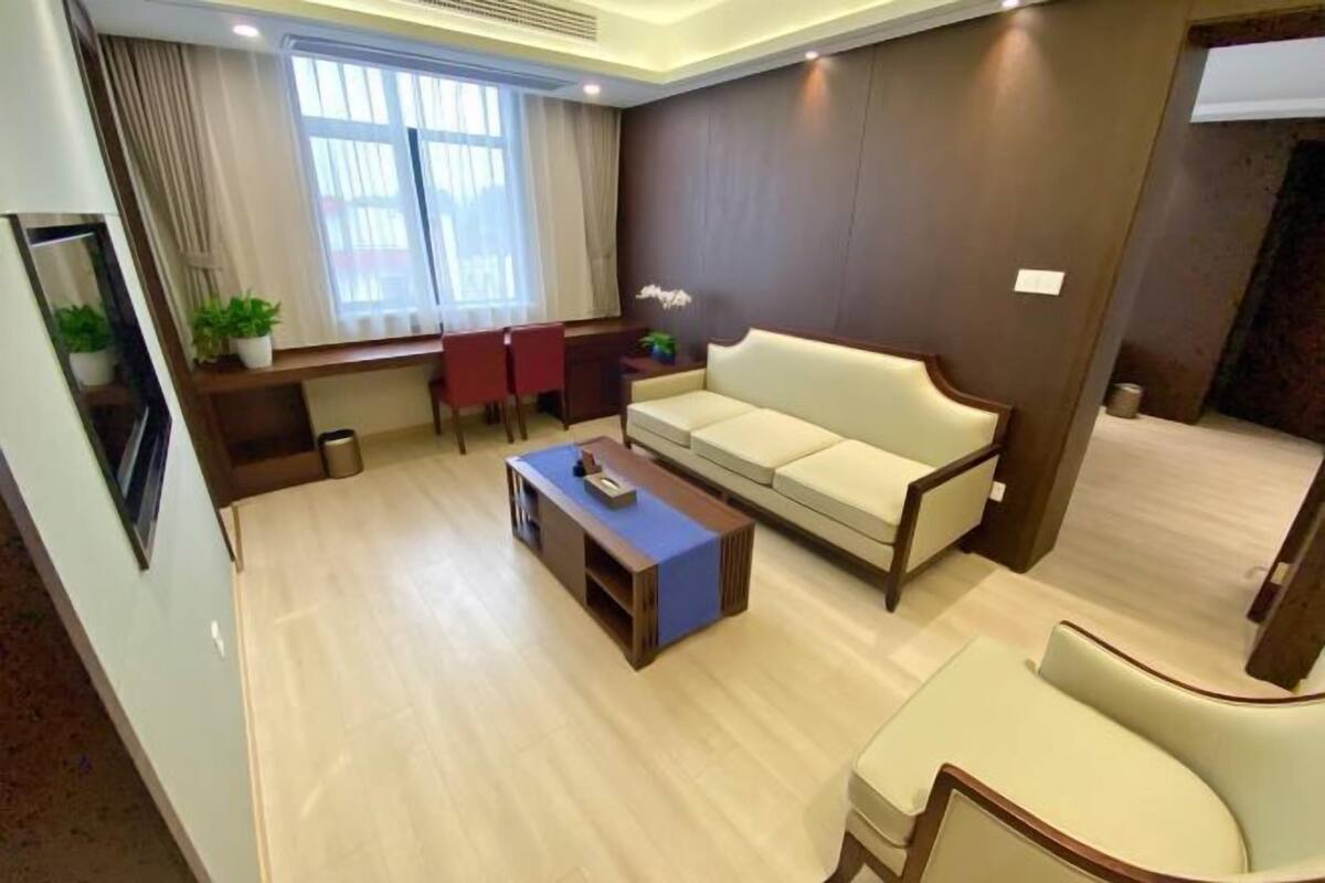 A modern living room with a sofa, coffee table, and chairs. There is a window with curtains, a small table with plants, and a wall-mounted shelf.