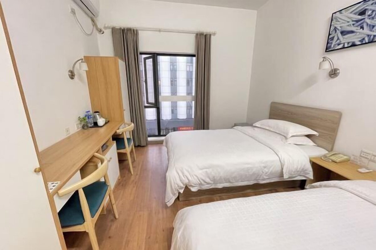 A hotel room with two beds, a desk, a chair, a window with curtains, and a wall-mounted phone.
