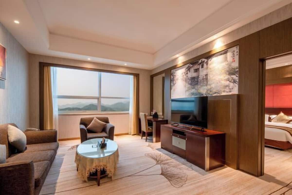 A modern hotel room with a large window, a sofa, a coffee table, a TV, and a bed.