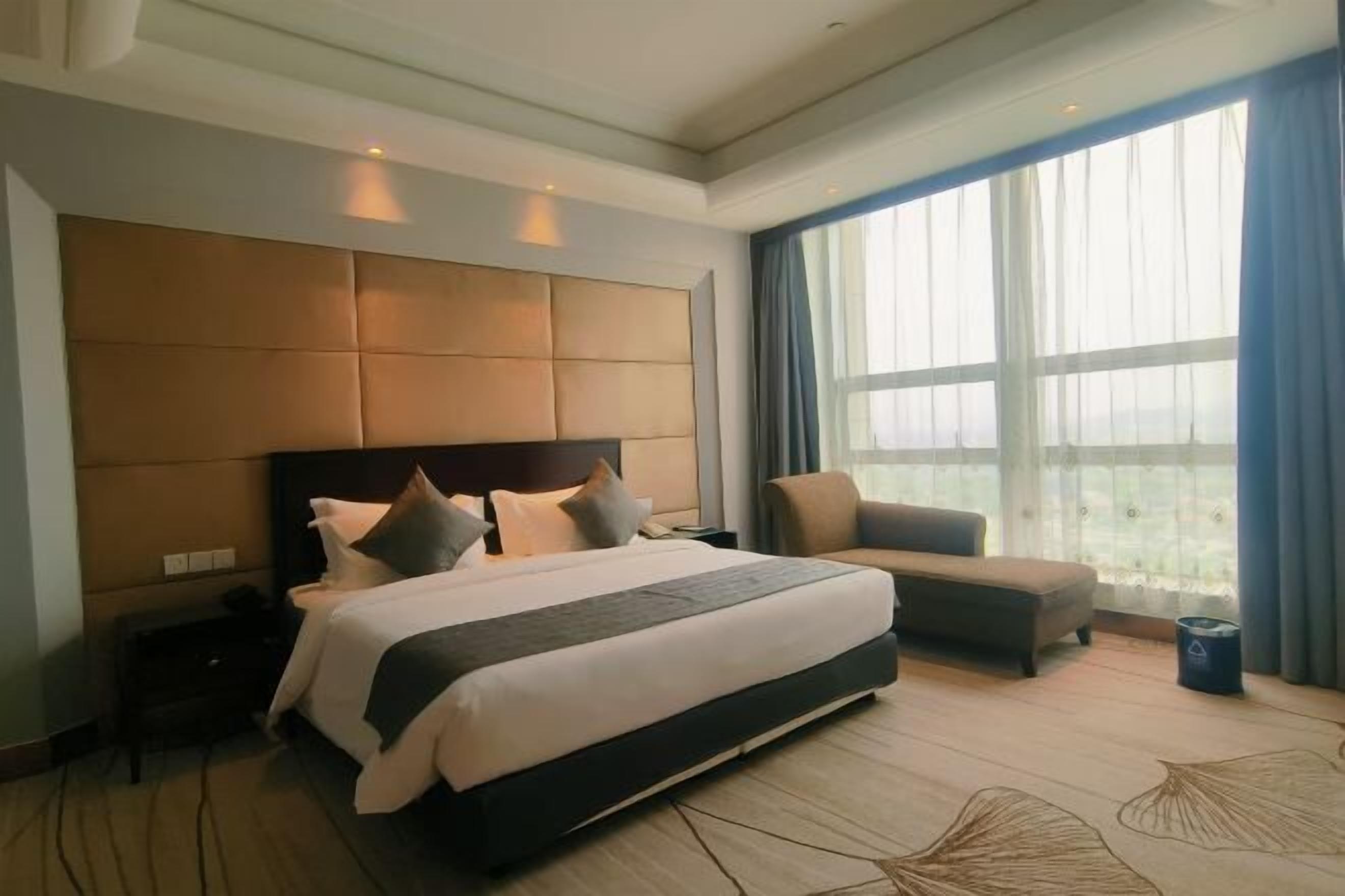 A modern hotel room with a large bed, a sofa, and a view of the city through large windows.
