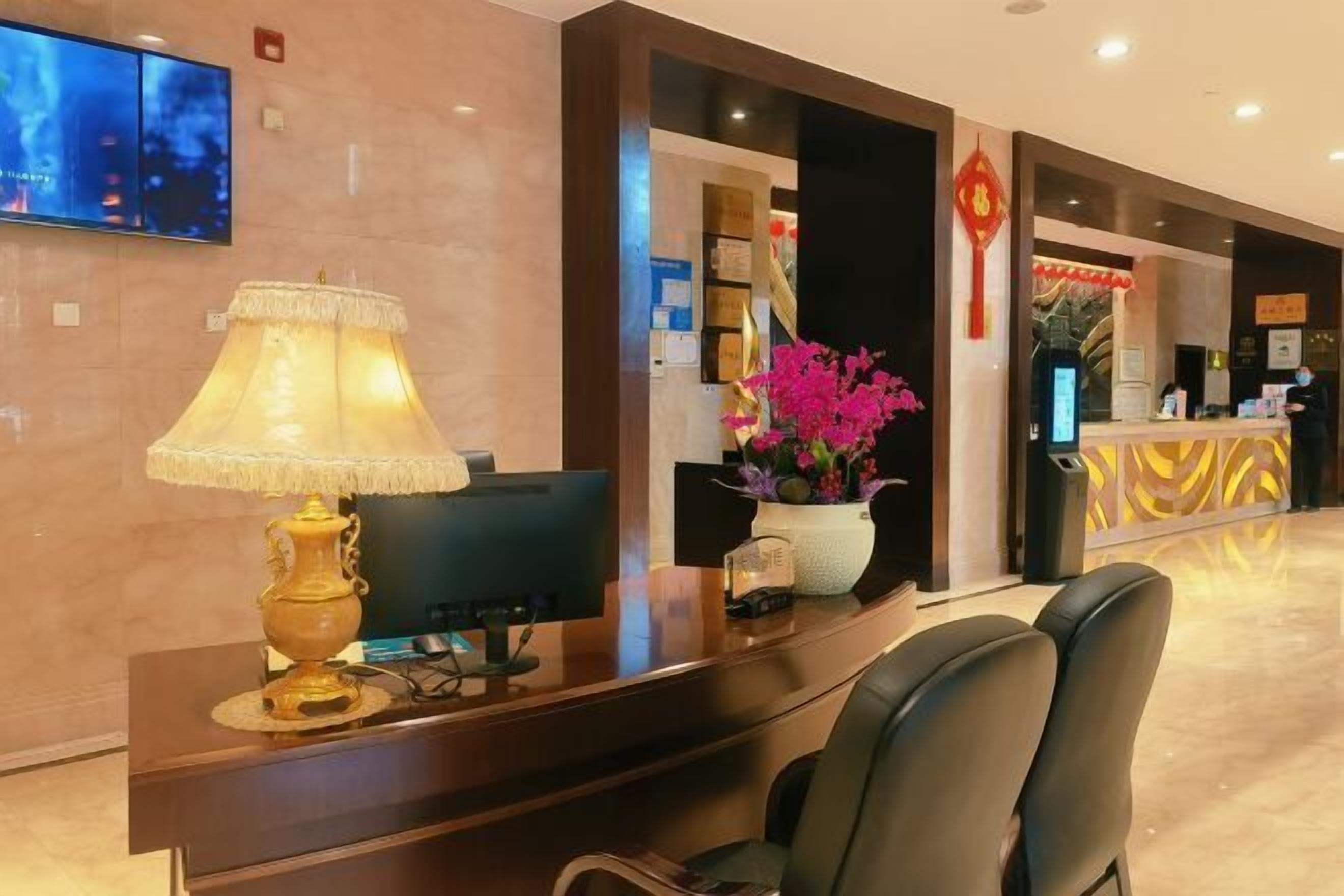 A hotel reception area with a desk, chairs, a television, a decorative lamp, and a vase with flowers.