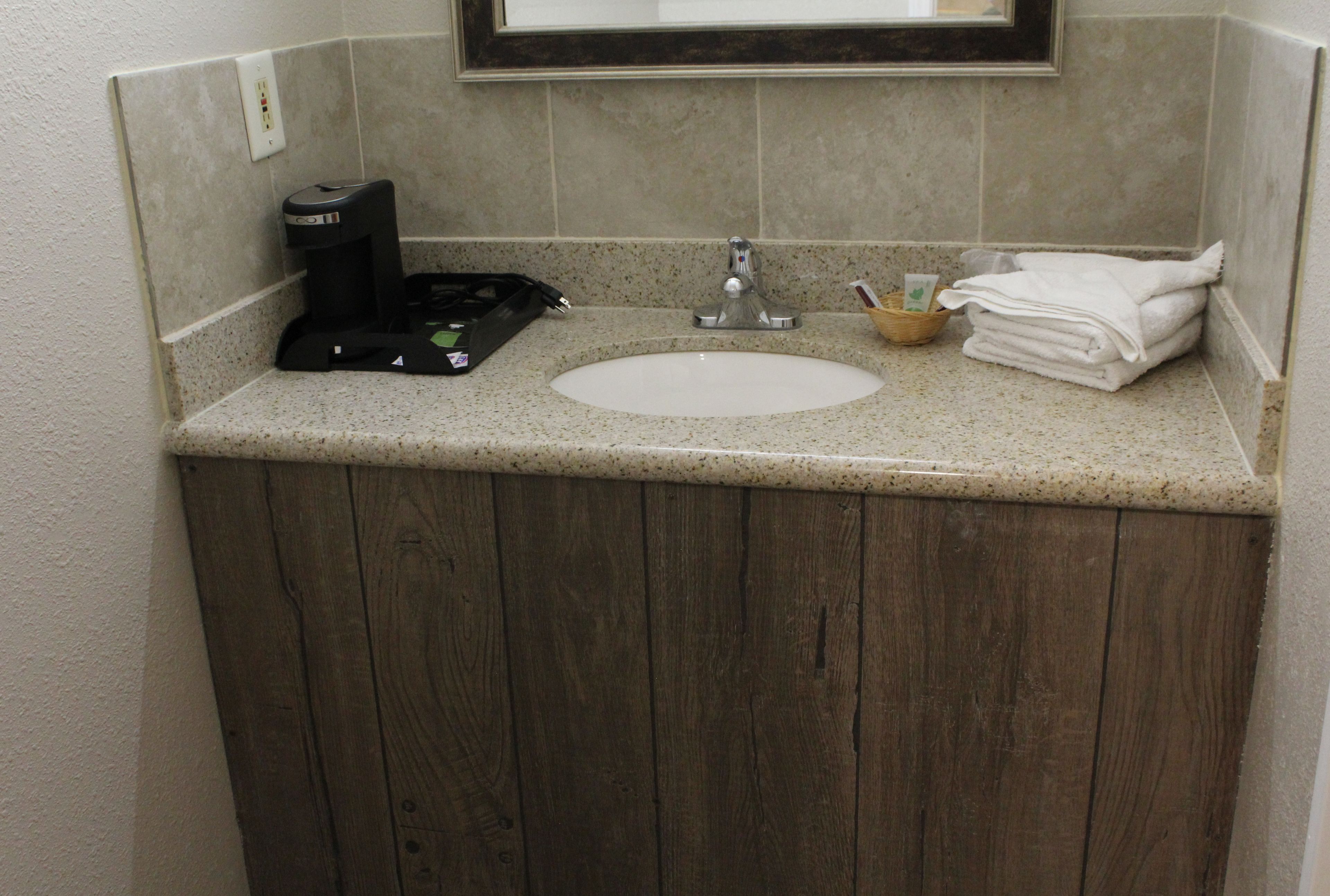A bathroom with a wooden vanity, a sink, a coffee maker, and a stack of white towels.