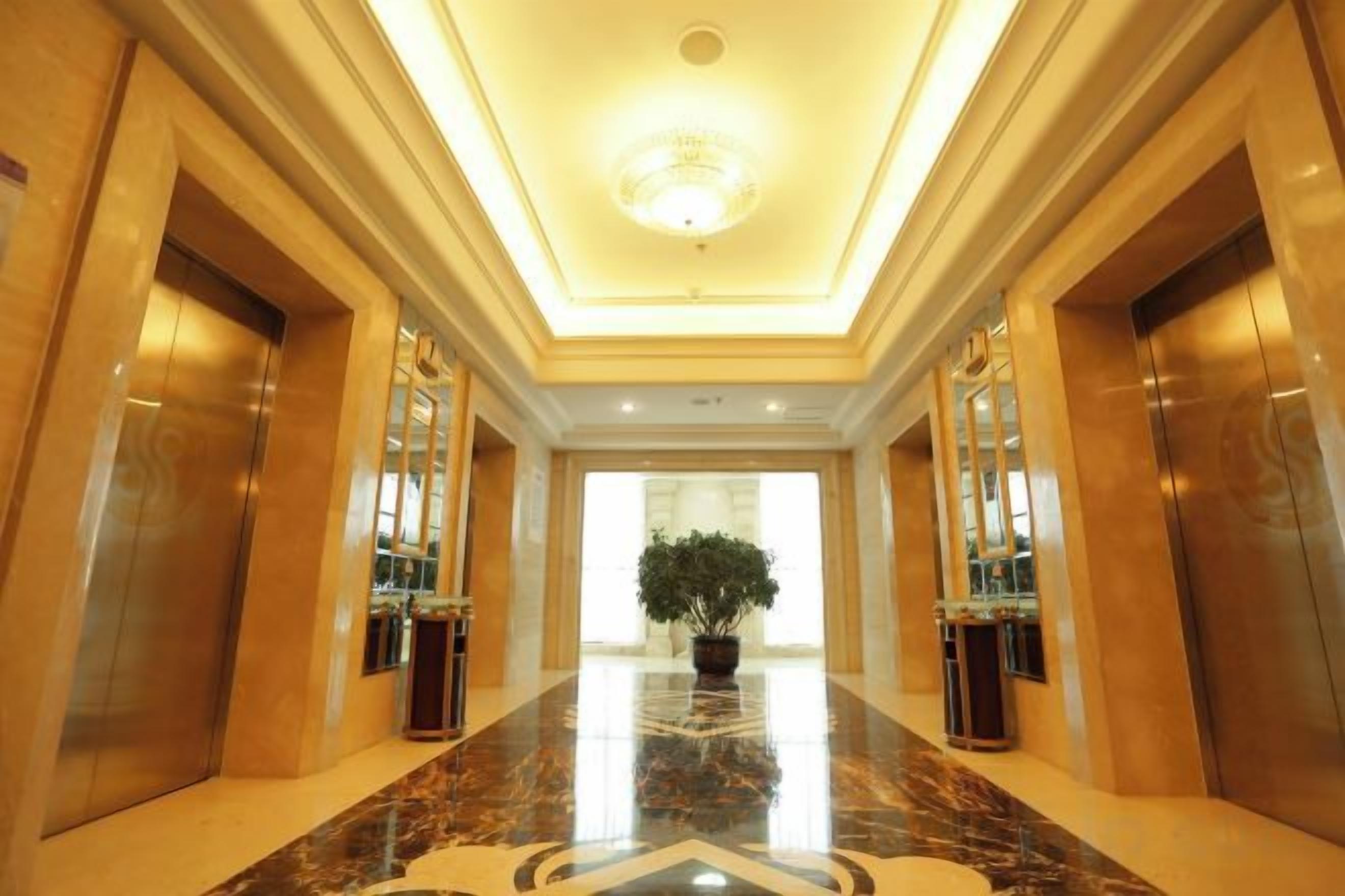 A hotel corridor with marble flooring, a central potted plant, and two elevators on either side.
