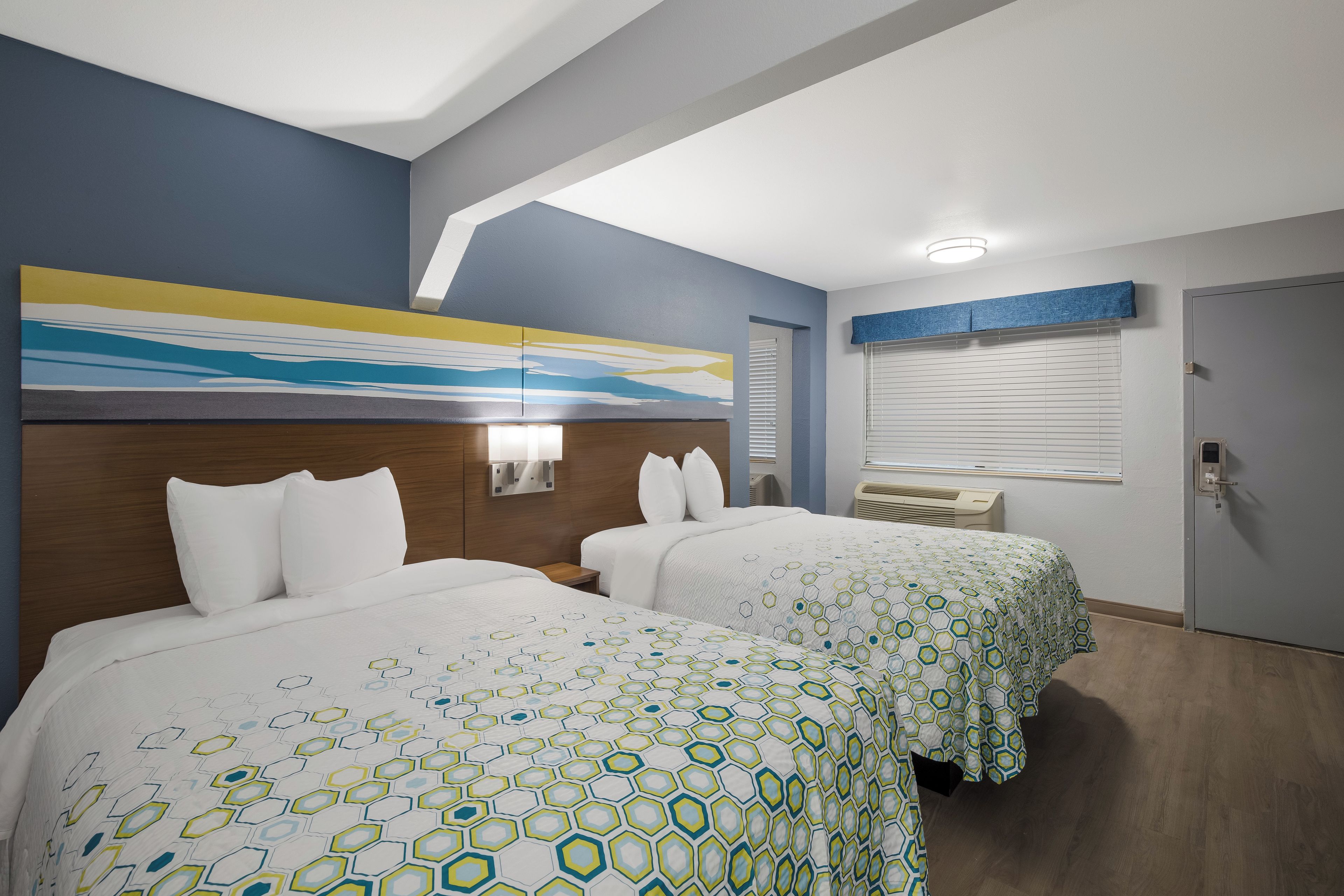 Une chambre d’hôtel avec deux lits, un grand tableau abstrait accroché au mur et une fenêtre avec des stores.