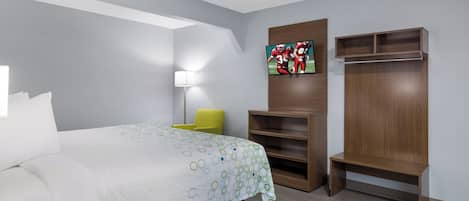 A hotel room with a bed, a TV mounted on the wall, a chair, and a wooden wardrobe.
