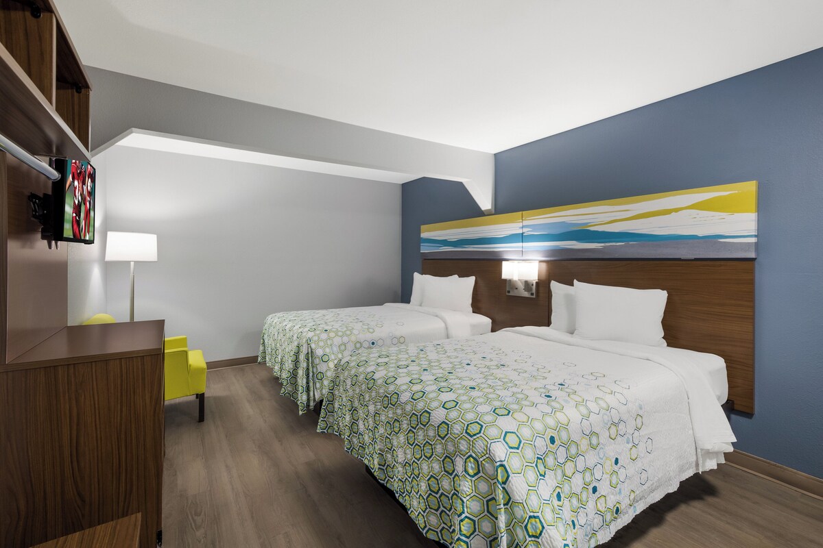 A hotel room with two beds, a desk, a chair, a painting on the wall, and a TV mounted on the wall.