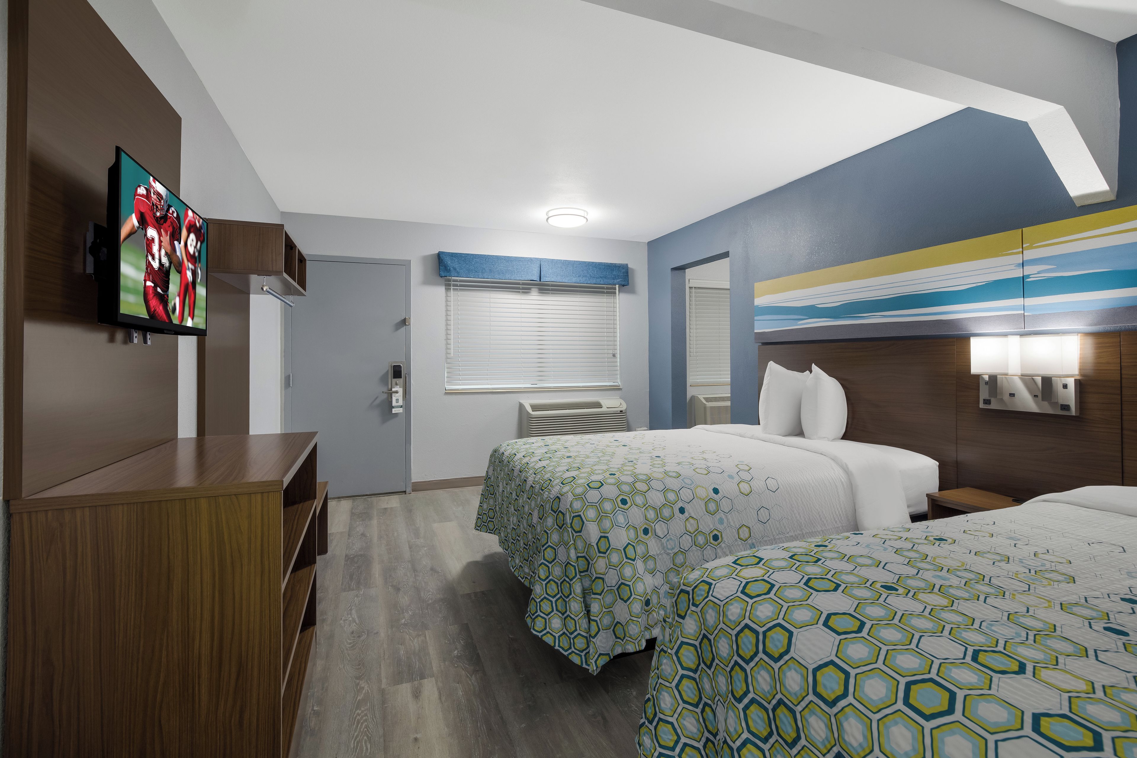 A hotel room with two beds, a TV, a desk, and a window with blinds.