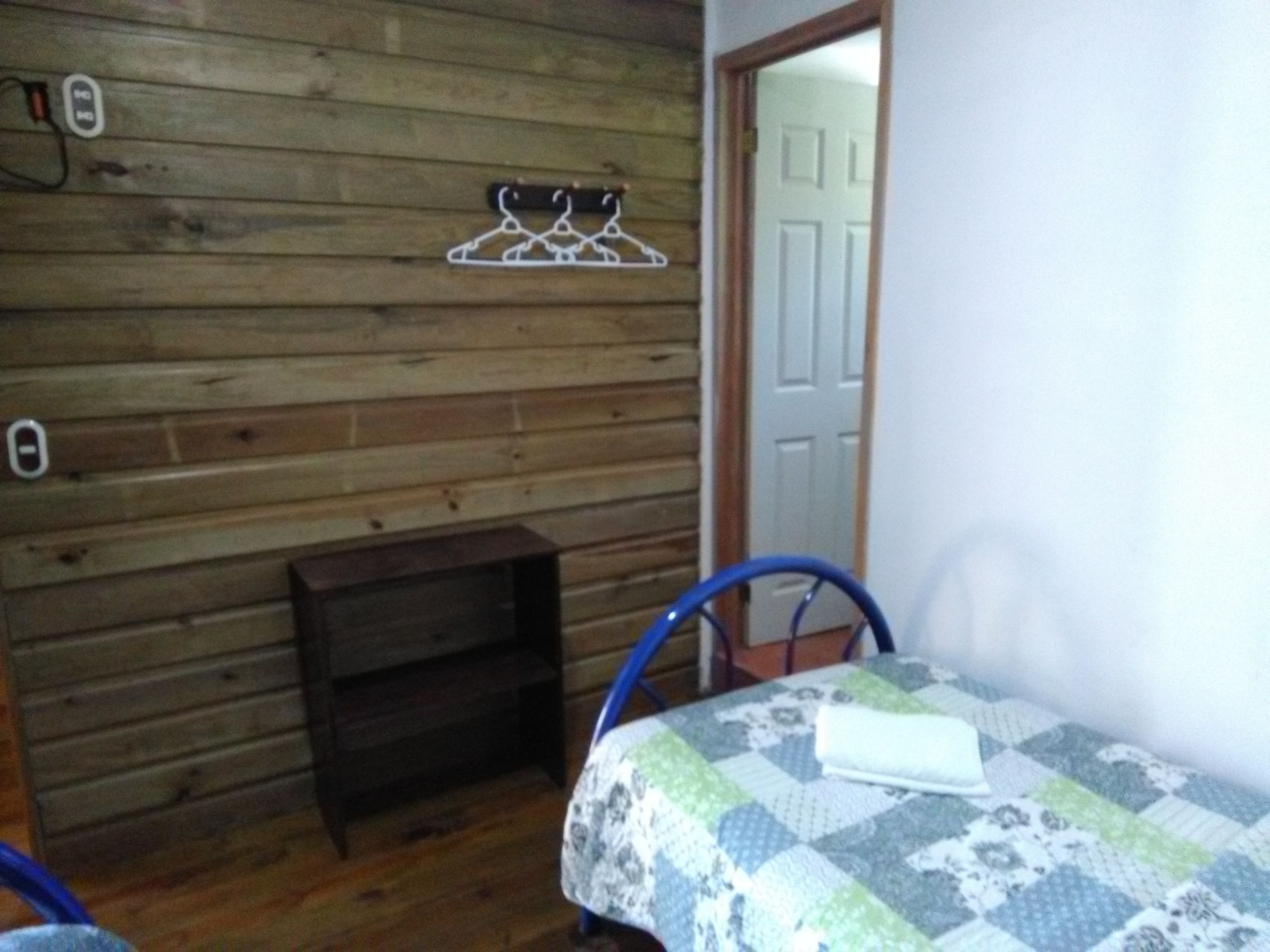 A small room with wooden walls, a bed with a patterned bedspread, a chair, and a small wooden shelf.