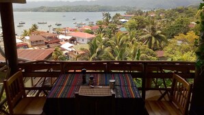 Vista desde un balcón sobre un pueblo costero con barcos en el agua, casas y palmeras.