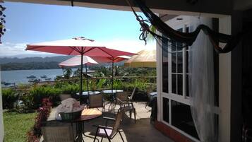 Een terras buiten met tafels en stoelen, rode parasols en uitzicht op een meer en bergen.
