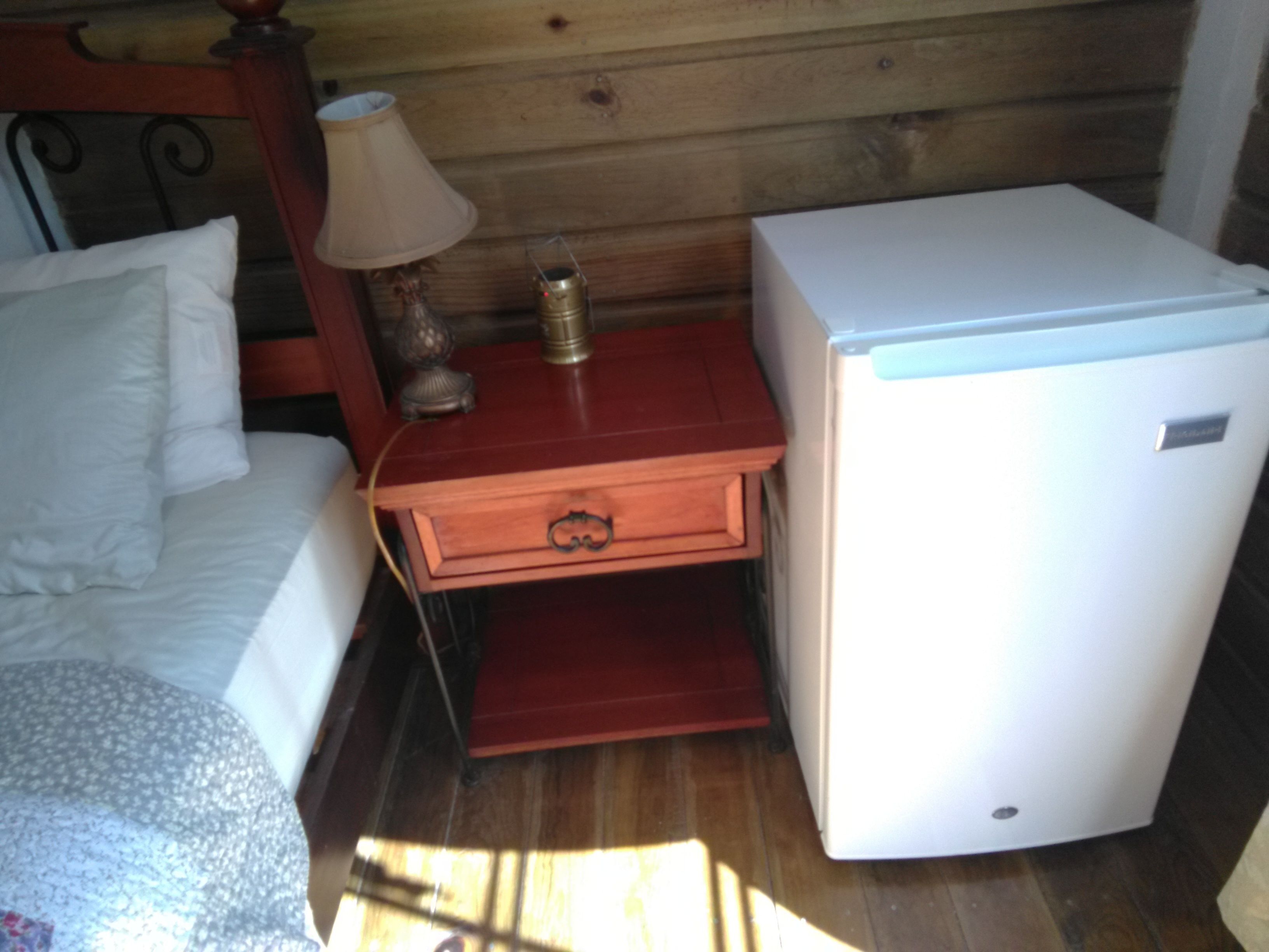 Una habitación pequeña con una cama, una mesita de noche con lámpara, una pequeña mesa y un refrigerador blanco.