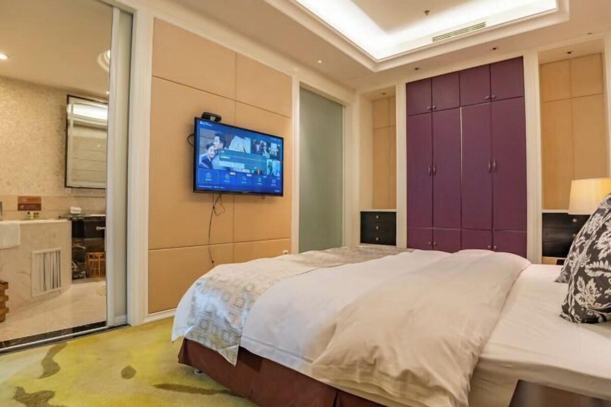 A hotel room with a bed, a television mounted on the wall, a wardrobe, and a small table with a lamp.