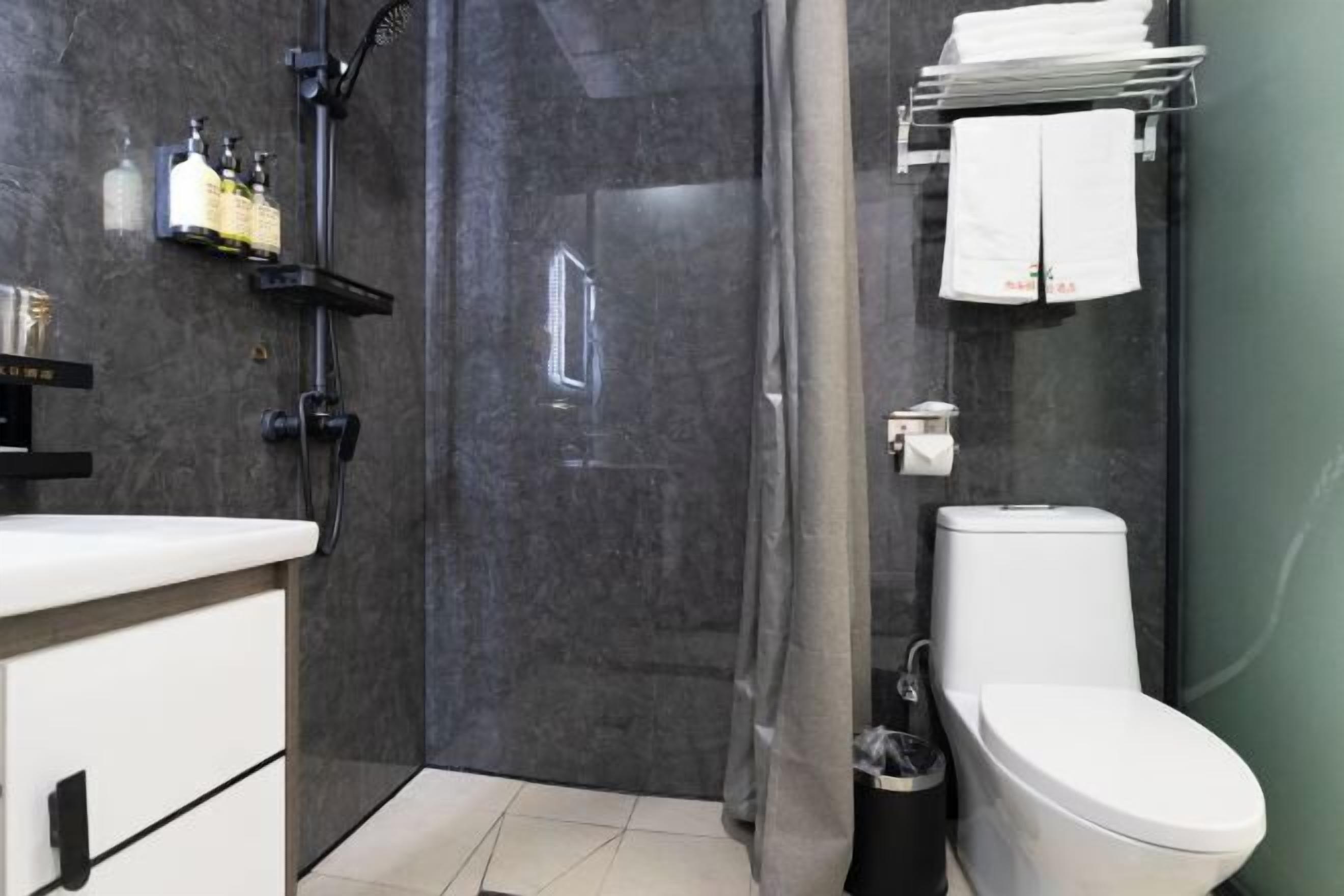 A modern bathroom with a shower, toilet, and sink. There are bottles on a shelf, a towel rack, and a trash bin.