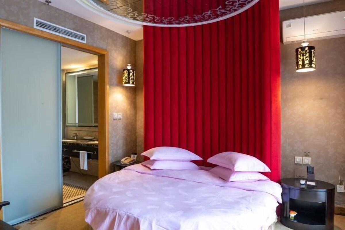 A hotel room with a large bed, a red curtain, a bedside table with a lamp, and a mirror.