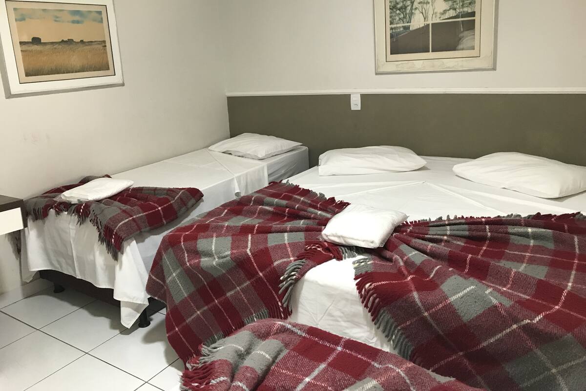 A room with two single beds, each with white linens and a red and grey plaid blanket. There are two framed pictures on the walls, one depicting a landscape and the other trees.