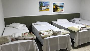 Three single beds are arranged in a row, each with a sheet, pillow, and folded blanket. Two framed paintings adorn the walls.