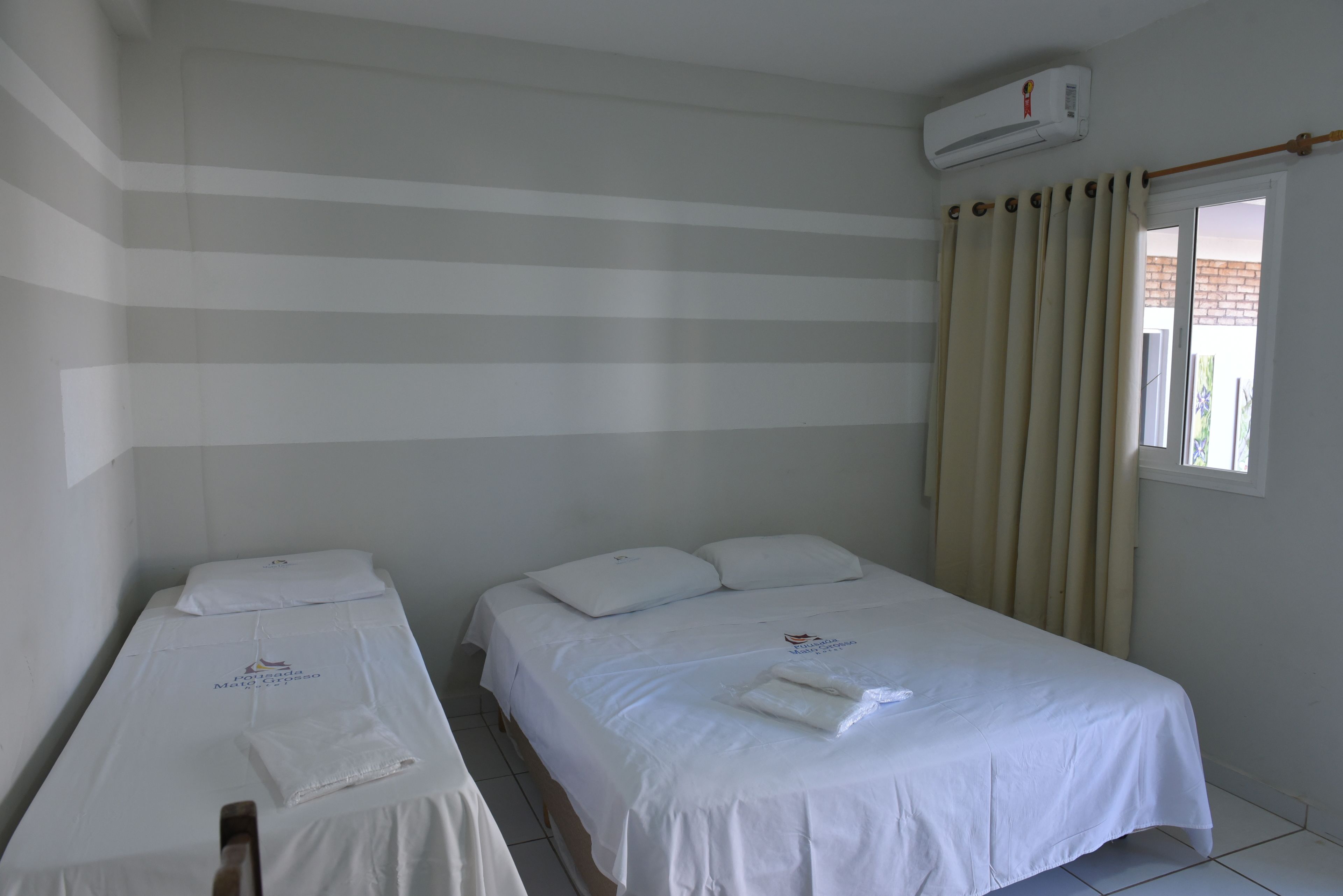 A hotel room with two single beds, a window with curtains, and an air conditioning unit on the wall.