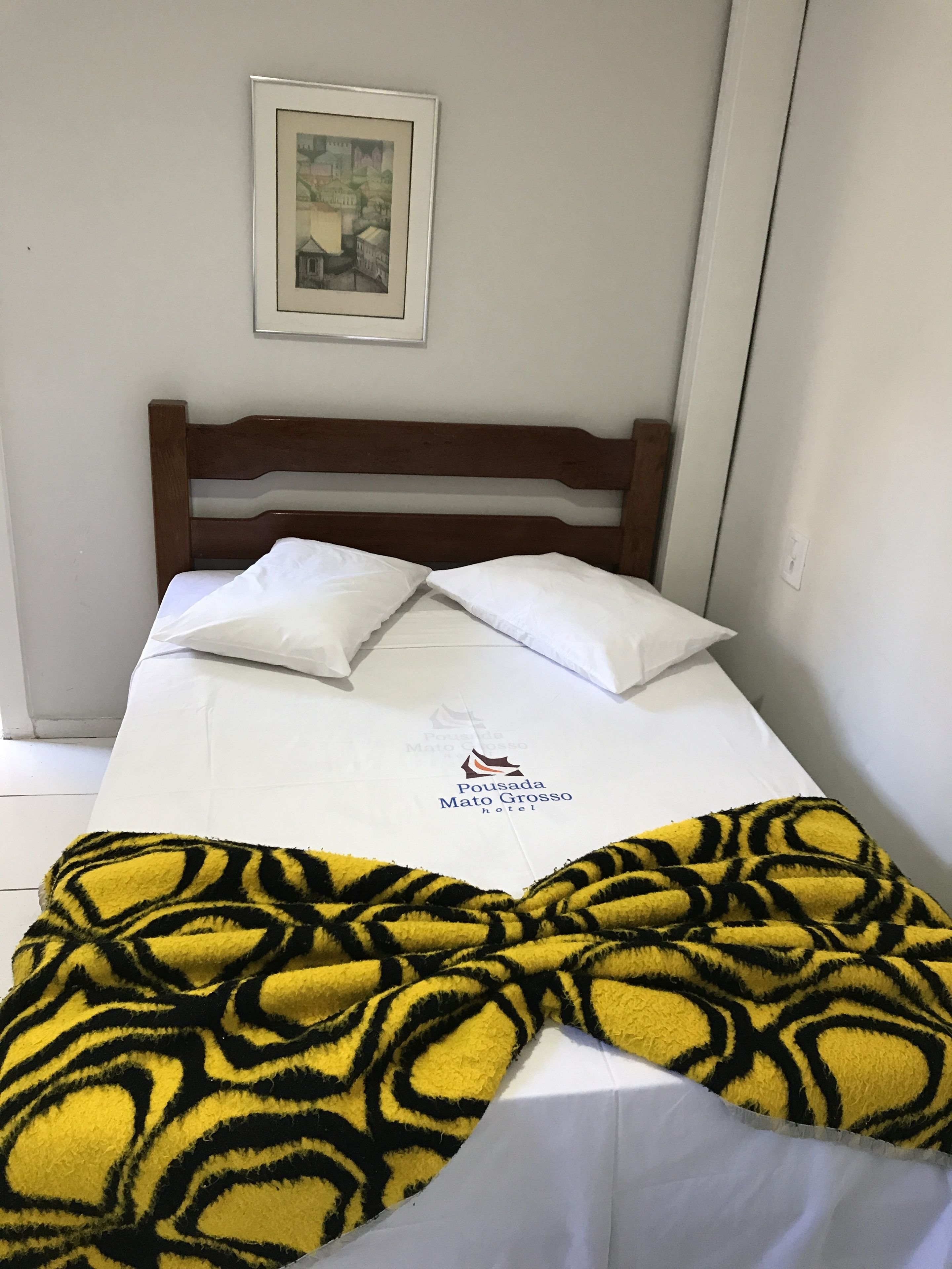 A bed with a white sheet, two pillows, a framed picture on the wall, and a yellow patterned blanket.