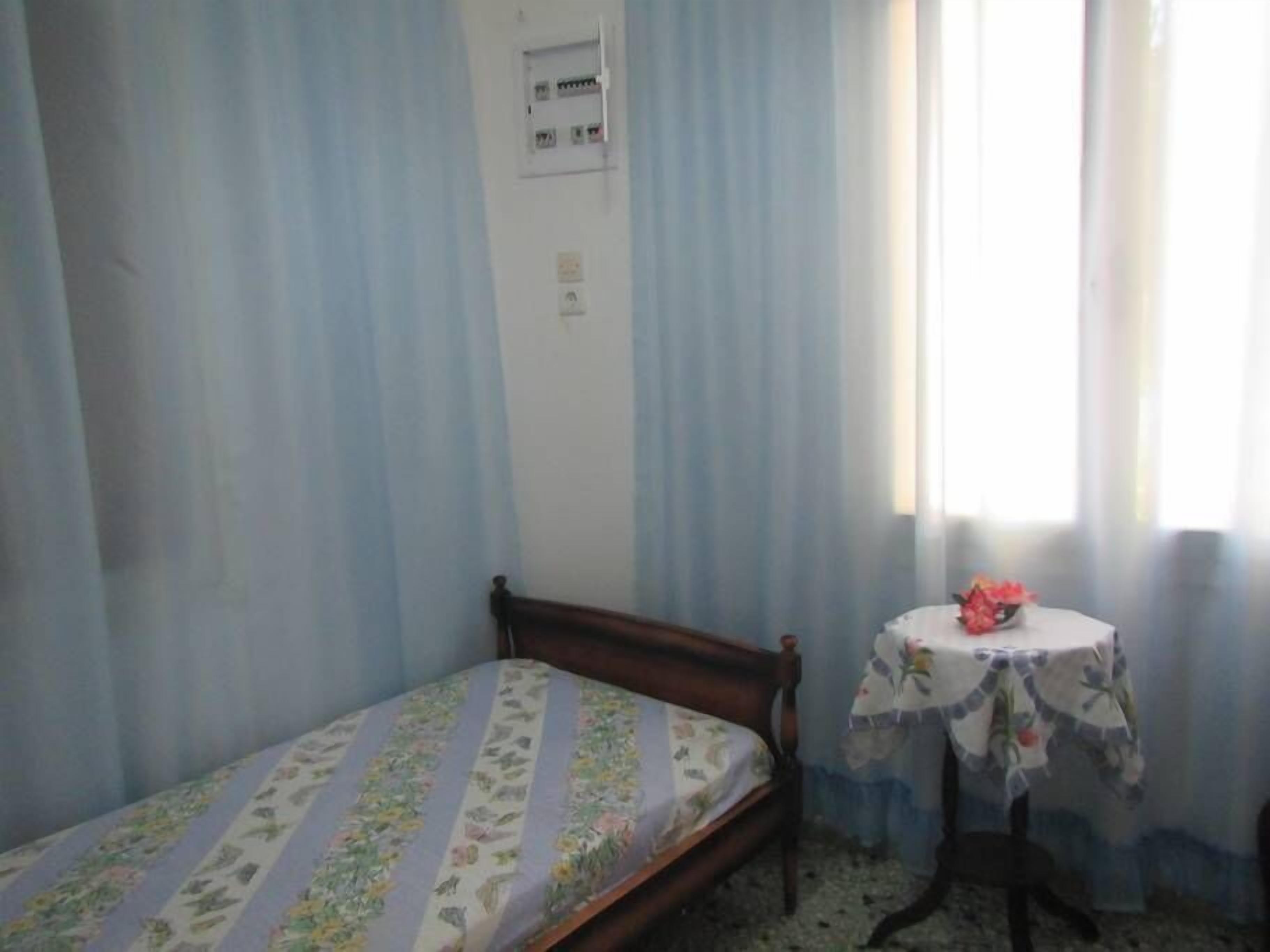 Un dormitorio con una cama individual, una mesita con mantel floreado y una ventana con cortinas azul claro.