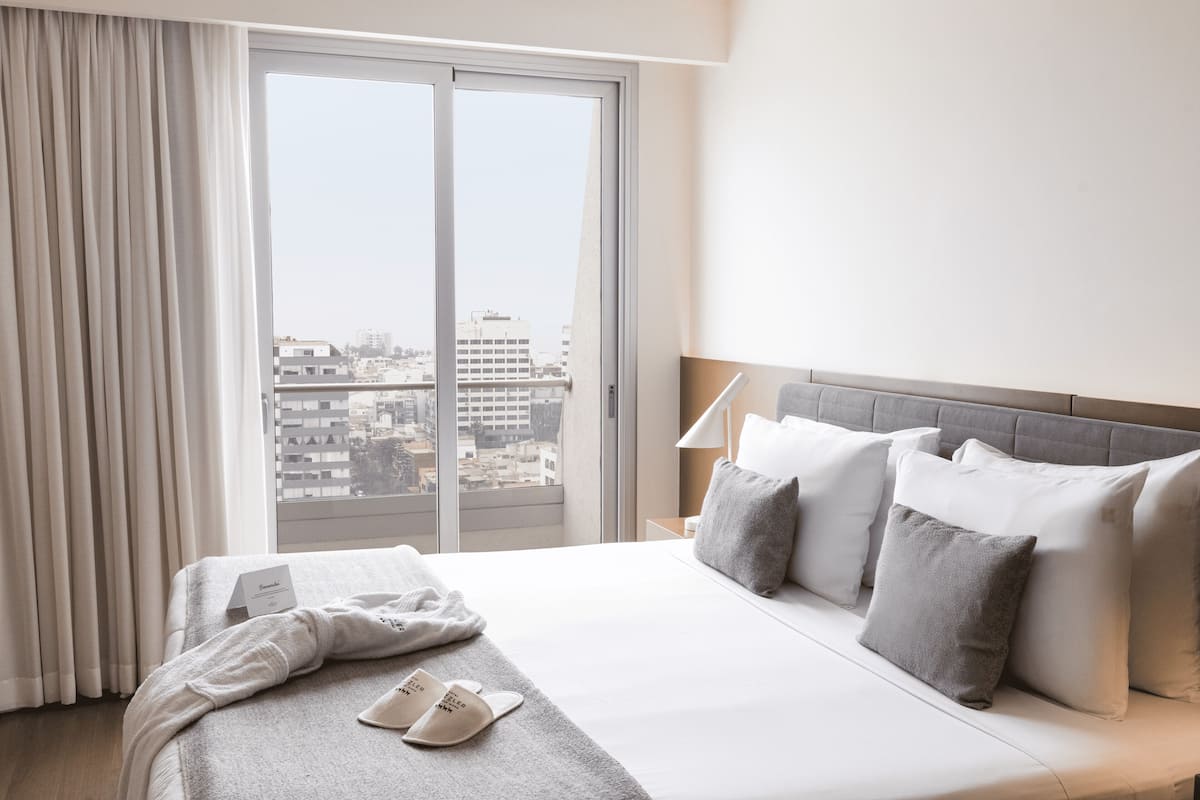 Ein Hotelzimmer mit einem großen Bett, grauer Bettwäsche, Blick auf die Stadt durch eine Schiebetür und einem Nachttisch mit Lampe.