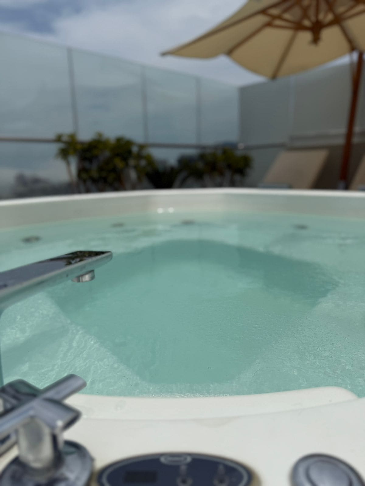 A hot tub with a control panel, surrounded by a glass enclosure with a view of the sky and some plants.
