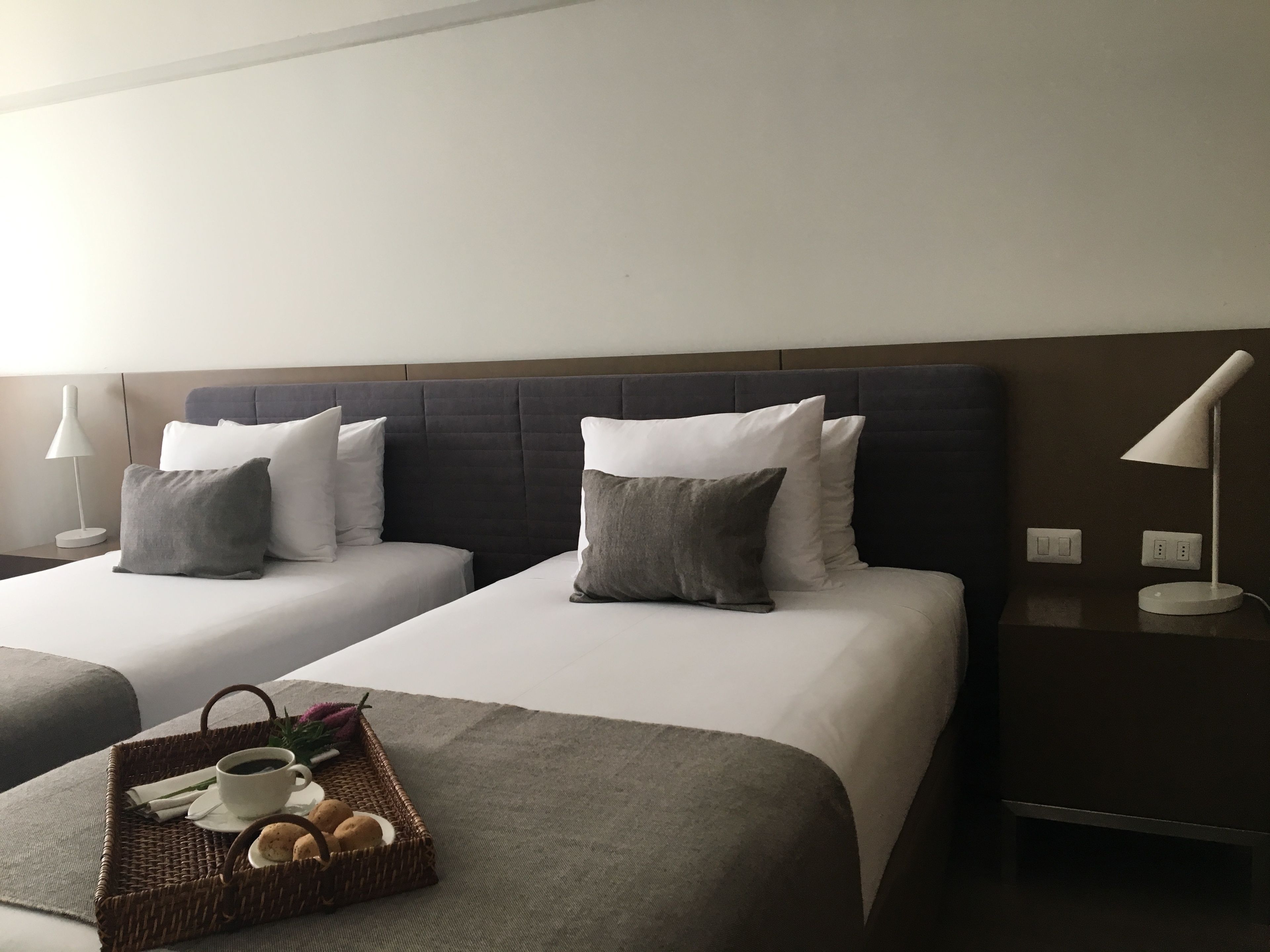 A hotel room with two beds, a nightstand, a lamp, and a tray with breakfast items.