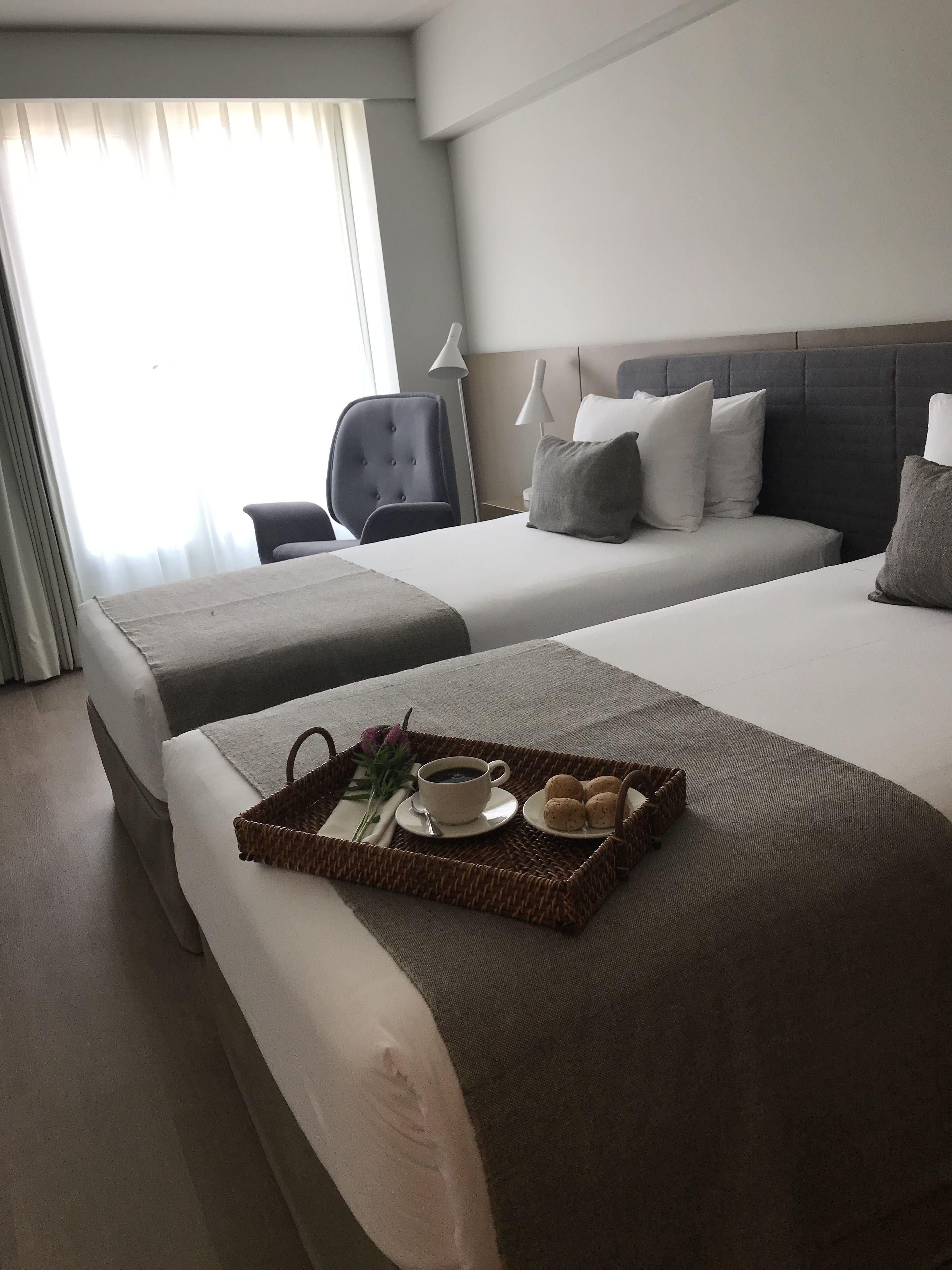 A hotel room with two beds, a desk, and a chair. There is a tray with a cup of coffee, a flower, and some pastries on the bed.