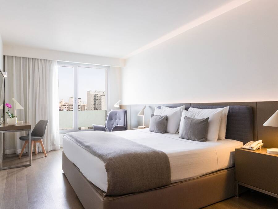 A modern hotel room with a large bed, a desk with a chair, a telephone, and a lamp.