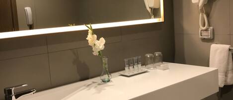Un baño moderno con tocador blanco, un espejo grande, un secador de pelo montado en la pared y un florero con flores en la encimera.