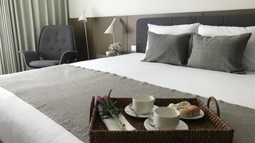 Una cama bien hecha con ropa de cama gris, una silla gris, una mesita de noche con una lámpara y una bandeja con cosas para el desayuno.