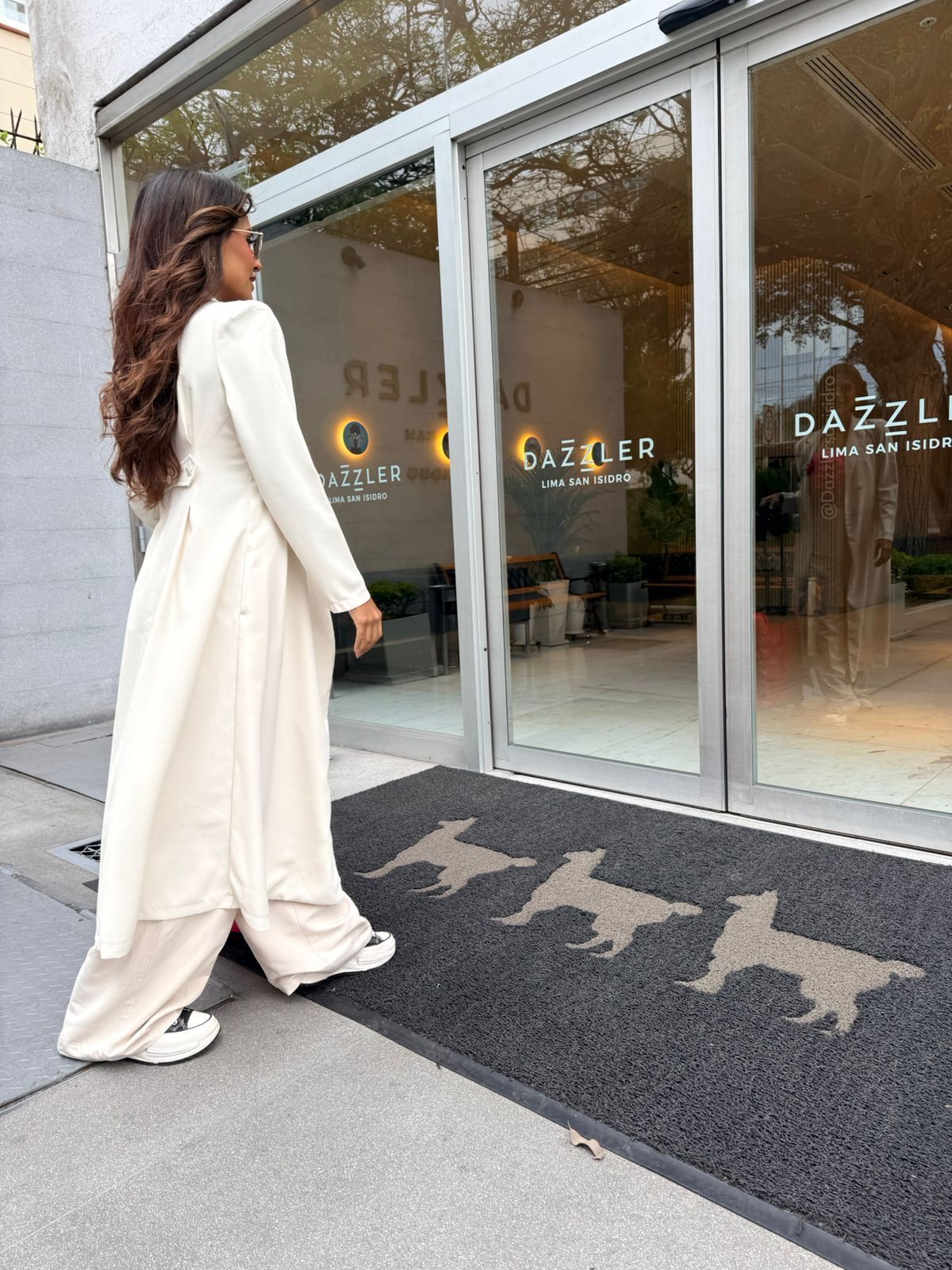 A woman in a white dress walking towards a building with large glass doors and a black doormat with animal silhouettes.