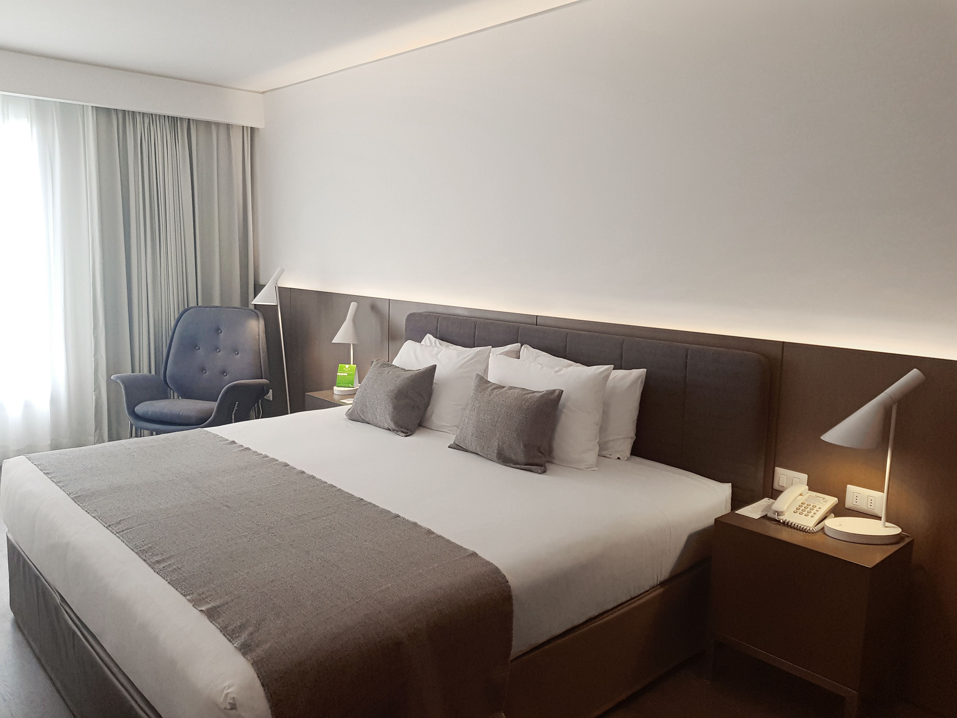 A hotel room with a large bed, a chair, a bedside table with a lamp, and a phone.