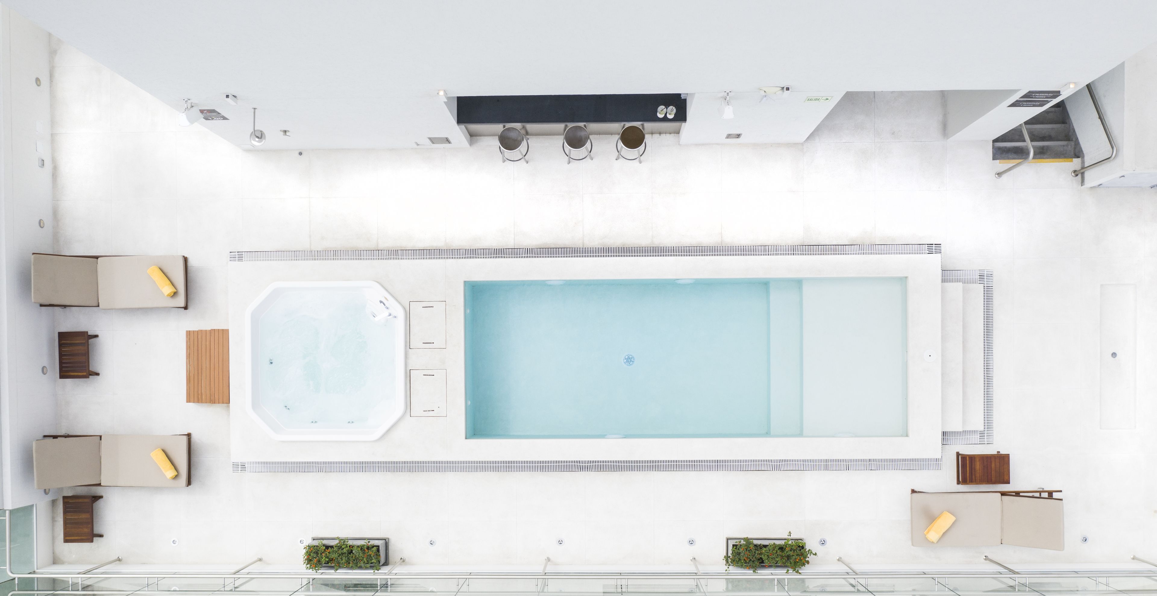 An aerial view of a modern indoor swimming pool area with a large rectangular pool, a smaller hot tub, and seating arrangements.